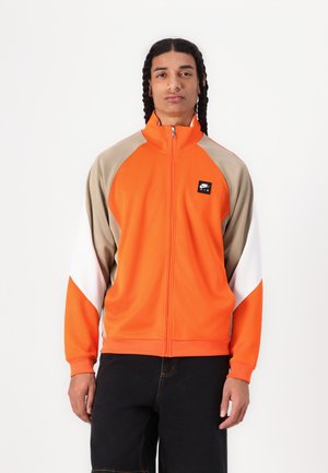 Veste zippée orange avec des manches raglan beige et blanches, ornée d'un logo Nike noir sur la poitrine. Fabriquée dans un matériau lisse et léger.