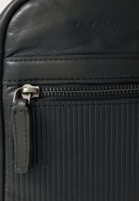 Seção de mochila preta em couro com ranhuras verticais texturizadas, um zíper de metal e uma etiqueta de couro lisa com gravação "bugatti".