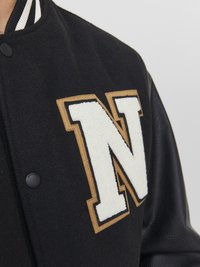 Zwarte varsityjas met leren mouwen en een groot wit "N"-embleem omlijnd met beige op de borst.