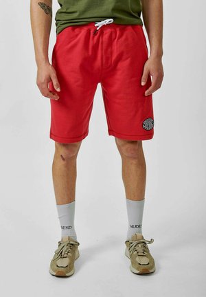 Shorts en coton rouge avec une taille élastique et un cordon blanc. Comprend des poches latérales et un écusson rond sur la jambe gauche.