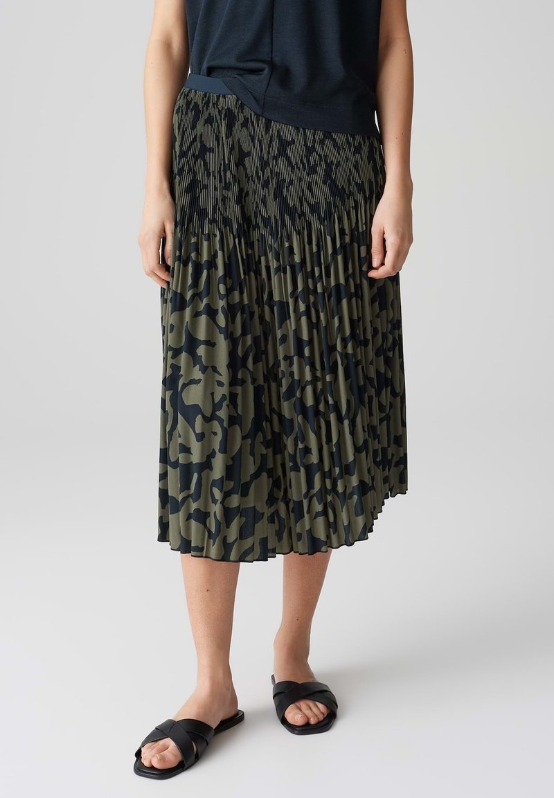 Jupe plissée vert foncé avec un motif abstrait noir audacieux, dotée d'une taille plate et d'une coupe fluide. Associée à des sandales noires.