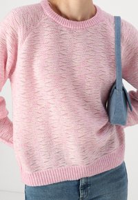 Osoba ubrana w jasnoróżowy sweter o teksturowanym splocie i niebieskie dżinsy, nosząca małą niebieską torbę na ramię po prawej stronie.