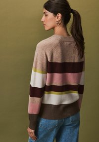Pull en tricot avec des rayures horizontales en beige, bordeaux, rose, blanc et olive. Présente un ourlet côtelé et un col rond. Associé à un jean bleu.