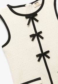 Beżowa sukienka bez rękawów o teksturowanym wykończeniu z czarnymi akcentami, trzy czarne kokardy z przodu oraz metka rozmiaru dla dzieci w wieku 6-7 lat.