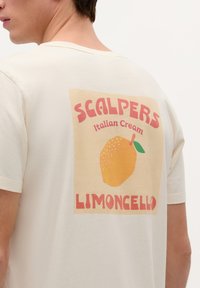 Kermanvärinen puuvillapaita, jossa on suuri graafinen kuvasto selkäpuolella. Kuvassa on sitruuna ja värikäs typografia, jossa lukee 'SCALPERS Italian Cream Limoncello'.