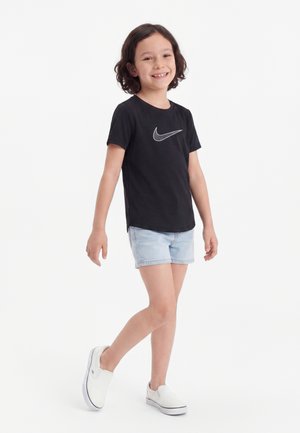 Smilende barn med bølget hår iført sort Nike t-shirt, lyse denimshorts og hvide slip-on sko, gående foran hvid baggrund.