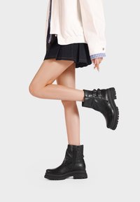 Stivali alla caviglia in pelle nera con punta arrotondata, doppie fibbie e finitura testurizzata. Suola spessa che offre aderenza e stabilità.