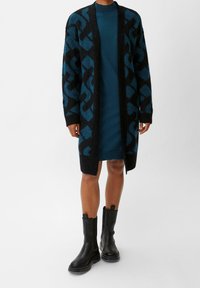 Cardigan nero e teal con motivo, realizzato in materiale a maglia morbido, design aperto sul davanti, abbinato a un vestito teal aderente e stivali neri alla caviglia.