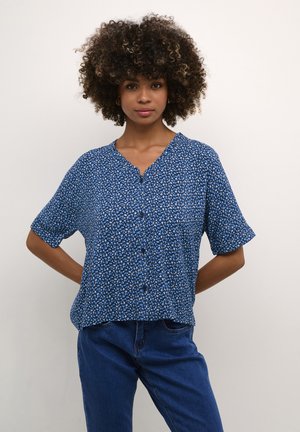 Chemise à boutons bleue à motifs floraux avec des manches courtes, dotée d'une coupe décontractée, d'un décolleté en V et d'un tissu léger. Portée avec un jean en denim.