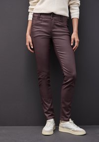 Bordeauxkleurige skinny broek van soepel materiaal, voorzien van zakken aan de voorkant en een dubbele knoopsluiting, gecombineerd met witte sneakers.