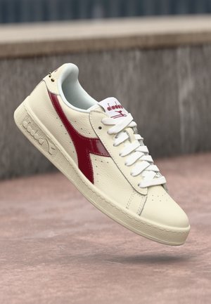 Cream leather sneaker with a glossy red accent, flat white laces, and textured rubber sole featuring a logo on the side and tongue.