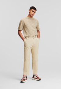 Beige mintázatú rövid ujjú ing, krémszínű nadr�ág oldalzsebekkel, és fekete-piros jegyekkel ellátott sneakers. A modell kezét a zsebében tartja.