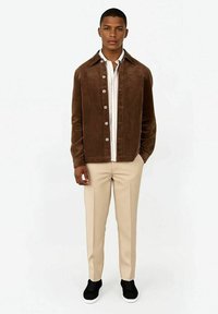 Bruine corduroyjas met kraag, knoopsluiting, gedragen over een wit shirt en lichtbeige broek. Zwarte sneakers maken de outfit compleet.