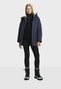 Marineblauwe pufferjack met een gevoerde capuchon van bont, gecombineerd met een zwarte gestructureerde trui en zwarte leggings. Model draagt zwarte sneeuwlaarzen.
