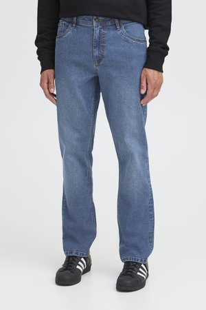 Man i raka blå jeans, svart sweatshirt och svarta sneakers med vita ränder som står på en enfärgad vit bakgrund.