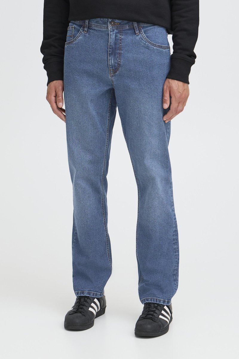 Bărbat purtând blugi drepti albaștri, un hanorac negru și adidași negri cu dungi albe, stând pe un fundal simplu alb.