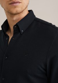 Črna srajca s patentnimi gumbi, izdelana iz teksturirane tkanine, z ovratnim ovratnikom in vezenim logotipom na prsih. Gumbi so temno modri.
