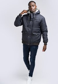 Svart vadderad jacka med huva, med grå foder, framknappar och två stora framfickor. Bärs med mörka jeans och vita sneakers.