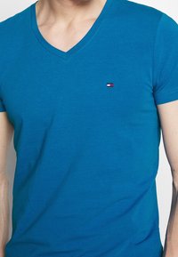 Blaues V-Ausschnitt-T-Shirt aus glattem Baumwollstoff. Mit einem kleinen gestickten Logo auf der linken Brust und kurzen Ärmeln.