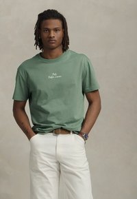 Mladic s dredovima nosi zeleni Polo Ralph Lauren majicu, bijele hlače, smeđi remen i sat na ruci, stojeći s rukama u džepovima.