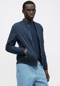 Junger Mann, der eine marineblaue Bomberjacke über einem dunkelblauen Hemd und hellblauen Jeans trägt, mit einer Hand in der Jackentasche, stehend vor einem weißen Hintergrund.