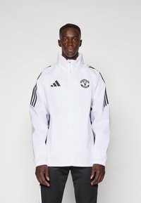 Hvid zip-jakke med høj krave, Adidas-logo, Manchester United-badge og sorte striber på ærmerne. Glat tekstur, atletisk pasform.