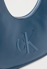 Μπλε δερμάτινη τσάντα χειρός με ανάγλυφο λογότυπο CK και εμφανές ασημένιο φερμουάρ κατά μήκος της πάνω άκρης, σε λευκό φόντο.