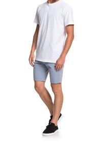 Muž oblečený v jednoduchém bílém tričku s krátkým rukávem, světlých šedých šortkách a černých teniskách, stojí s váhou na jedné noze na bílém pozadí.