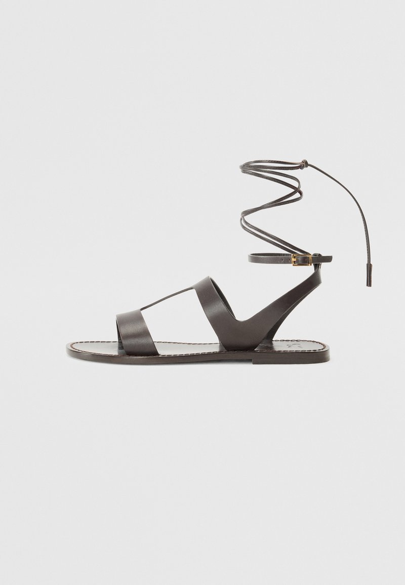 Sandal de cuero marrón con suela plana, que cuenta con un diseño minimalista, tira de tobillo curva y múltiples cortes para transpirabilidad.