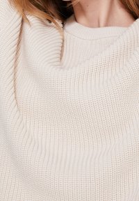 Osoba v krémovém svetru s hrubým pletením, má klasický kulatý výstřih a dlouhé rukávy, odkrytou část krku a vlasů.