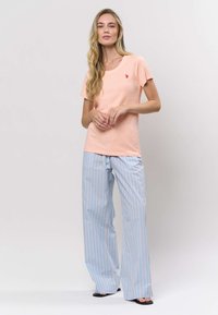 Vaalean persikanvärinen T-paita, jossa on pieni punainen logo, yhdistettynä sinisiin raidallisiin leveälahkeisiin housuihin. Asu on rento ja siinä on pehmeä materiaali.