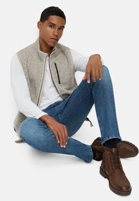 Grijze fleece vest met witte mouwen, ritszak, blauwe spijkerbroek en bruine veterschoenen. Zittende pose op een witte achtergrond.