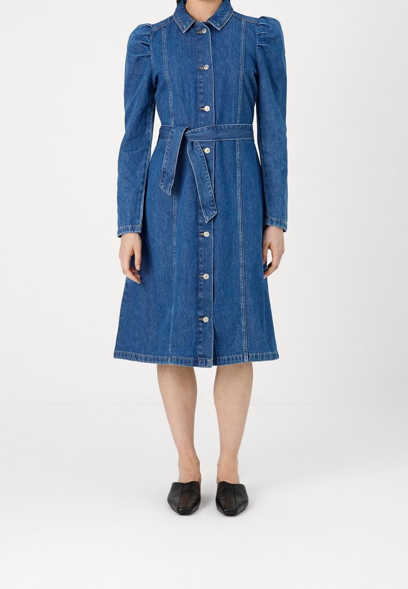 Femme portant une robe en jean bleu longueur genoux avec des manches longues bouffantes, des boutons sur le devant, une ceinture à la taille et des ballerines noires pointues.
