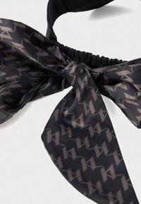 Czarna opaska na głowę z elastycznym tyłem i przymocowaną kokardą, z powtarzającym się szarym wzorem geometrycznym na gładkim materiale.