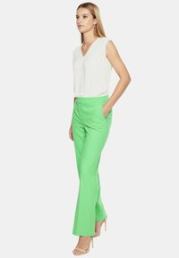 Zielone spodnie z rozszerzanymi nogawkami, z kieszeniami i zamkiem błyskawicznym z przodu, w połączeniu z białą bluzką bez rękawów. Materiał jest gładki i lekki.