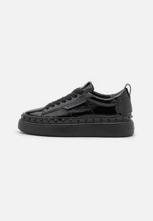 Czarny lakierowany sneaker z grubą podeszwą, ozdobą z ćwiekami wzdłuż śródpodeszwy, czarnymi sznurówkami i pętelką na pięcie, widok z boku.