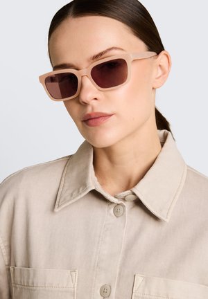 Femme aux cheveux foncés tirés en arrière portant une chemise beige oversize boutonnée et des lunettes de soleil rectangulaires rose clair avec des verres foncés.