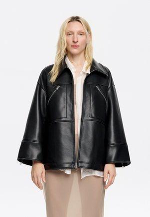 Black faux leather oversized jacket with zippers, two chest pockets, wide collar, and a smooth texture, worn over a light-colored top.