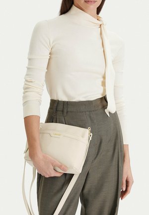 Donna che indossa un top a maniche lunghe color crema con collo a fiocco e pantaloni grigi a vita alta, che tiene una borsa a tracolla color crema con dettagli dorati.