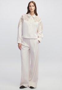 Licht crèmekleurige blouse met kantdetails op de schouders, knoopsluiting aan de voorkant en een losse pasvorm. Gecombineerd met bijpassende wijde broek in een vergelijkbare tint.