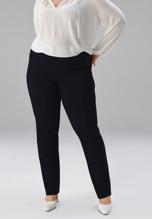 Frau trägt schwarze, eng anliegende Hose, weiße langärmlige Bluse und weiße Pumps, steht mit einer Hand in der Hüfte vor einfarbigem Hintergrund.