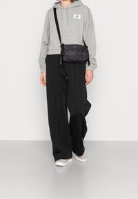 Sudadera gris con puños elásticos, bolsa negra con diseño texturizado, pantalones holgados negros y zapatillas ligeras grises, de pie sobre un suelo gris.