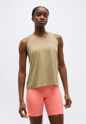 Mujer con camiseta sin mangas beige y pantalones cortos de ciclismo color rosa coral, de pie contra un fondo claro y liso.