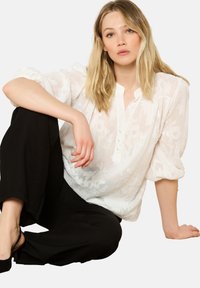 Blouse blanche à motifs floraux avec une patte de boutonnage et des manches légèrement ballons, assortie à un pantalon noir ample. Texture lisse.