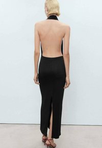 Vestido negro de cuello halter con espalda descubierta, diseño ajustado y abertura lateral, hecho de una tela suave. La modelo lleva sandalias rojas con tiras.