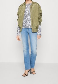 Oliwkowa satynowa kurtka bomberka na szarym topie w panterkę, zestawiona z jasnoniebieskimi jeansami o prostych nogawkach i czarnymi, wiązanymi szpilkami.