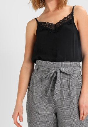 Kobieta ubrana w czarny top z koronkową lamówką i wysokie szare spodnie w kratę z paskiem wiązanym w talii, stojąca na jednolitym tle.