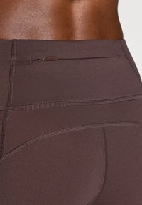 Barna sportleggings sima textúrával, kis cipzáras zsebbel és kontrasztos varrásokkal a varratok mentén.