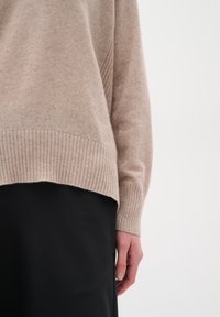 Beiger, weicher Strickpullover mit gerippten Ärmelbündchen und Saum. Das Design zeigt ein dezentes, strukturiertes Paneel an der Seite, kombiniert mit einem schwarzen Rock.
