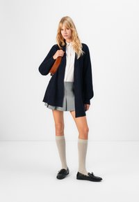 Jeune femme portant un cardigan noir, une blouse blanche, une jupe plissée grise, des chaussettes beiges montantes et des mocassins noirs, tenant un sac à épaule marron.